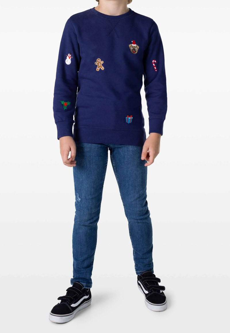 Dziecko w granatowym dresie z łatkami o tematyce świątecznej, w niebieskich dżinsach i czarnych trampkach na rzepy, stojące na białym tle.