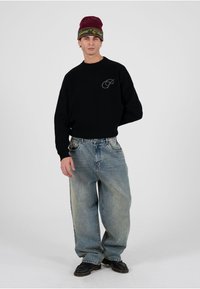 Sudadera negra con cuello redondo y logo bordado, combinada con vaqueros azules desteñidos y holgados, zapatos negros con cordones, rematado con un gorro de lana en tonos burdeos y verde.