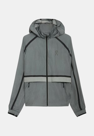 Jachetă ușoară gri deschis cu glugă, fermoar negru, accente negre și reflectorizante, manșete elastice și buzunare laterale cu fermoar.