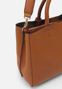 Sac cabas en cuir texturé marron avec une fermeture éclair dorée, une garniture noire contrastante, des poignées doubles et une bandoulière ajustable.