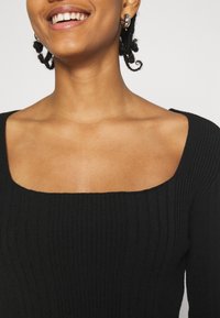 Haut noir à manches longues côtelé avec un large col carré et une texture douce, présentant des lignes horizontales accentuées tout au long.