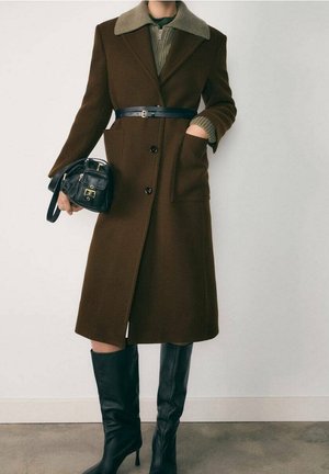 Manteau en laine marron avec un col beige, ceinturé à la taille. Il présente une coupe droite, deux poches avant, et des bottes en cuir noir montant jusqu'aux genoux.