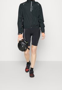 Chaqueta de ciclismo negra ligera con rayas reflectantes, combinada con pantalones cortos acolchados negros y zapatos de ciclismo negros con acentos rojos, sosteniendo un casco negro.