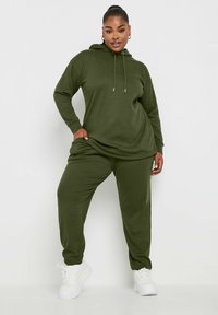 Felpa con cappuccio verde oliva e pantaloni abbinati in tessuto a costine. Presenta cordini e una vestibilità comoda, abbinati a scarpe da ginnastica bianche.