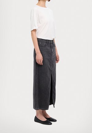 Osoba ubrana w białą koszulę z krótkim rękawem, czarną spódnicę maxi z denimu o wysokim stanie z przednim rozcięciem oraz czarne płaskie buty, stojąca bokiem.