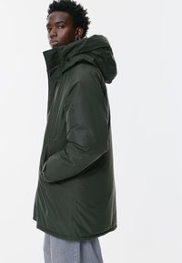 Geacă puffer verde închis cu guler înalt și glugă ajustabilă, având o textură netedă și buzunare laterale. Siluetă clasică, alungită.