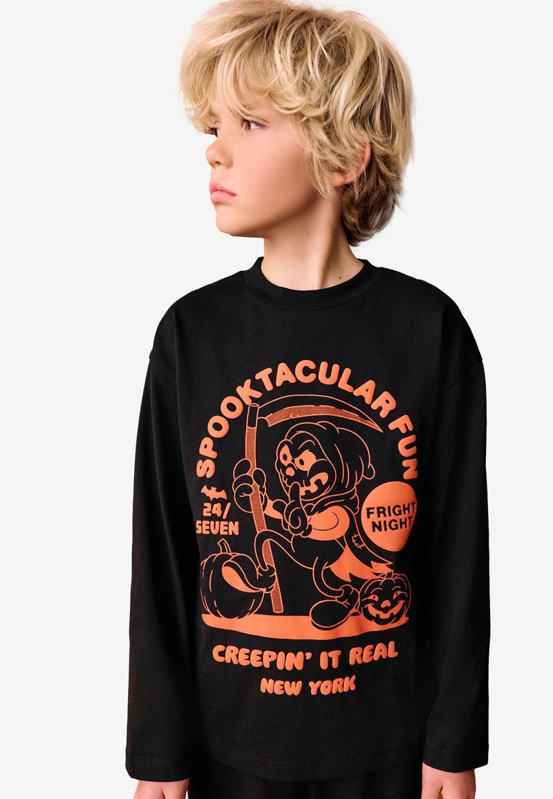 Chemise noire à manches longues avec un motif graphique orange vif incluant un personnage de dessin animé, des citrouilles, et le texte : « Spooktacular Fun ». Coupe décontractée.