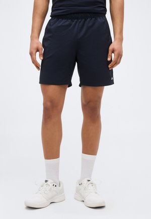 Άνδρας που φοράει μαύρο αθλητικό σορτς, λευκές κάλτσες μέχρι τον αστράγαλο και λευκά αθλητικά παπούτσια, στέκεται μπροστά σε απλό ανοιχτόχρωμο φόντο.