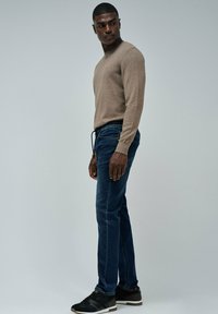 Hombre vistiendo un suéter beige, jeans oscuros con cordón y zapatillas deportivas negras, de pie y mirando hacia su izquierda contra un fondo liso.