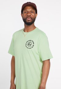 Camiseta de algodón verde claro con mangas cortas y un escote redondo, que presenta un logotipo gráfico negro central. Sin patrones ni acentos visibles.
