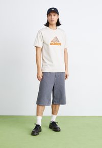 Tricou din bumbac bej cu logo-ul Adidas portocaliu, pantaloni scurți din denim gri, încălțăminte neagră, șosete albe și o șapcă bleumarin. O ținută simplă și lejeră.