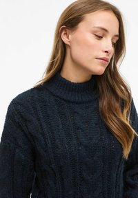 Kobieta z długimi brązowymi włosami, ubrana w ciemnogranatowy, gruby sweter z wysokim kołnierzem, patrząca w dół i nieco na bok.