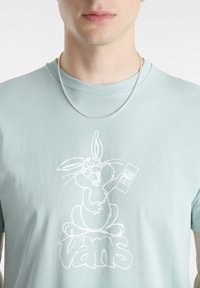 Camiseta de algodón azul claro con un gráfico de un conejo de dibujos animados blanco sosteniendo un control remoto, con el logo de "Vans" debajo. Manga corta y cuello redondo.