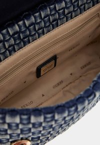 Intérieur d'un sac à main tissé bleu avec doublure beige, comporte une poche zippée et une étiquette noire affichant "GUESS" en or. Texture lisse.