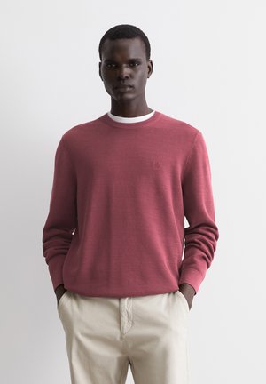Roter Strickpullover mit strukturiertem Muster, Rundhalsausschnitt, gerippten Bündchen und einem weißen Kragenakzent. Getragen mit hellbeigen Hosen.