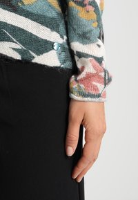 Stickad tröja med ett flerfärgat blommönster i grönt, rosa och gult. Inkluderar en blå detaljknapp och bärs över ett svart plagg.