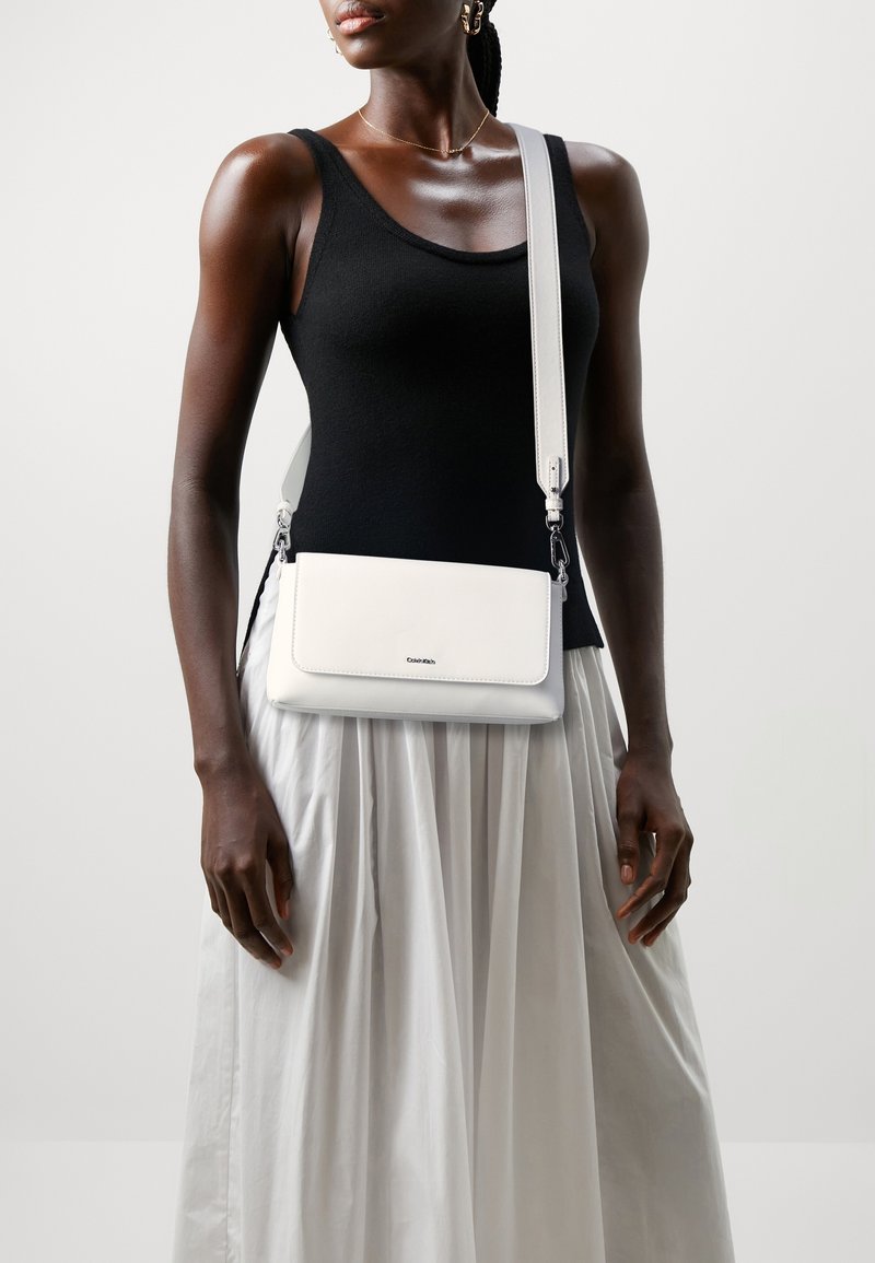 Femme portant un débardeur noir et une jupe plissée blanche, portant un sac bandoulière blanc Calvin Klein avec une large sangle.