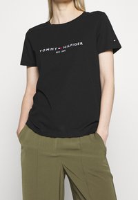 Žena v černém tričku Tommy Hilfiger s logem a olivovými kalhotami, s rukama v kapsách, na jednotném pozadí.