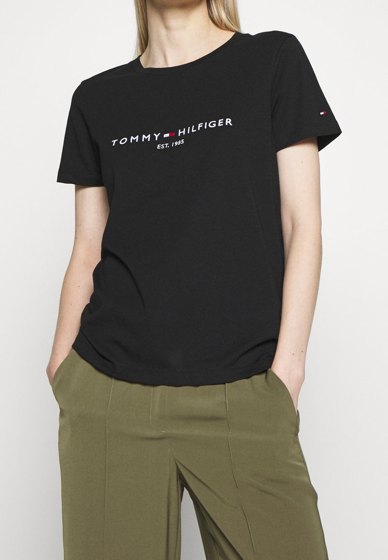 Žena v černém tričku Tommy Hilfiger s logem a olivovými kalhotami, s rukama v kapsách, na jednotném pozadí.