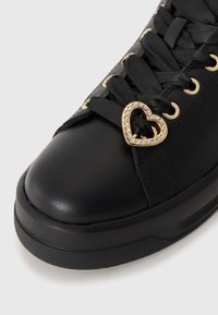 Black synthetic sneaker with a smooth texture, featuring gold hardware and a heart-shaped accent adorned with clear rhinestones.