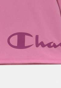 Capo in cotone rosa con un logo viola prominente che presenta un emblema circolare e il testo "Champion" in un carattere grassetto e fluido.