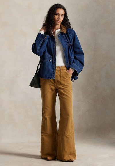 Jeans jakna z rjavim kordurjevim ovratnikom, kombinirana s svetlo rjavimi razširjenimi hlačami. Modelka drži majhno črno torbico in stoji na nevtralnem ozadju.