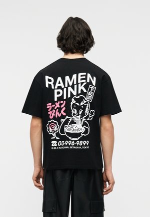 Person wearing black T-shirt with large white and pink graphic of cartoon eating ramen, Japanese text, and Tokyo address on the back.