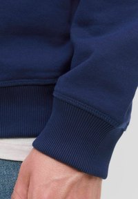 Prim-plan al încheieturii unei persoane care poartă un hanorac bleu închis cu manșete cu ţesătură rib și o cămașă albă, împreună cu blugi albaștri.