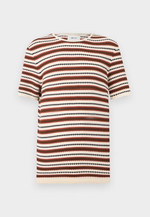 Ung mand stående med hænderne i lommerne iført en stribet kortærmet skjorte, løse beige bukser og brune åbne sandaler.