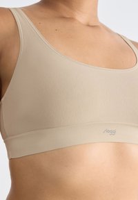 Beige bh med en mjuk, elastisk textur och bred kant. Har en rundad halsringning och tunna axelremmar. Logotyp tryckt på kanten.