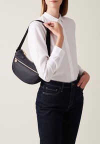 Piccola borsa a tracolla nera in pelle con finitura testurizzata, zip dorate e una lunga tracolla, indossata sopra una camicia bianca con colletto e jeans scuri.