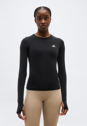 Femme portant un haut noir ajusté à manches longues avec des ouvertures pour les pouces et un legging beige, debout devant un arrière-plan clair uni.