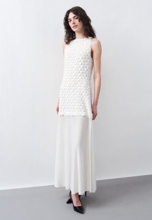 Γυναίκα με σγουρά σκουρόχρωμα μαλλιά, που φορά ένα λευκό αμάνικο τοπ με υφή και μία μακριά λευκή φούστα, στέκεται μπροστά σε ένα απλό φωτεινό φόντο.