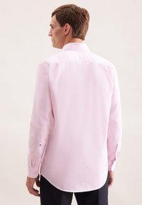 Uomo che indossa una camicia da uomo a maniche lunghe di un leggero colore rosa visto da dietro su uno sfondo semplice e chiaro.