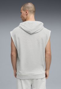 Άνδρας με κοντά μαλλιά, που φοράει αμάνικη γκρι ανοιχτόχρωμη μπλούζα με κουκούλα και αντίστοιχα σορτς, στέκεται με την πλάτη στραμμένη προς την κάμερα μπροστά σε γκρι φόντο.