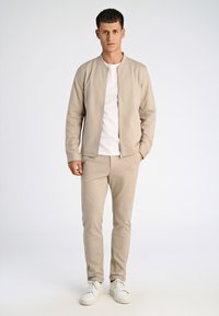 Man die een beige jack met rits draagt, beige bijpassende broek, wit T-shirt en witte sneakers, staand tegen een effen lichte achtergrond.