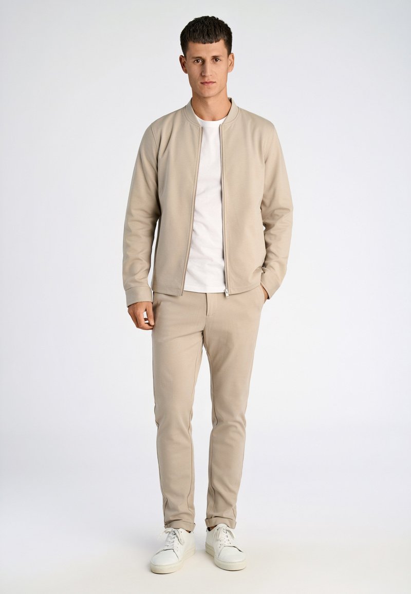 Man die een beige jack met rits draagt, beige bijpassende broek, wit T-shirt en witte sneakers, staand tegen een effen lichte achtergrond.