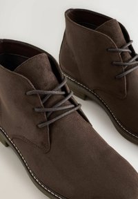 Botas de ante marrón hasta el tobillo con cordones grises, con un tacón bajo y costuras visibles, exhibidas sobre un fondo claro.