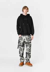 Sudadera negra con bolsillo frontal, combinada con pantalones sueltos de patrón gris y negro. Completan el look unos zapatos marrones con suela gruesa.