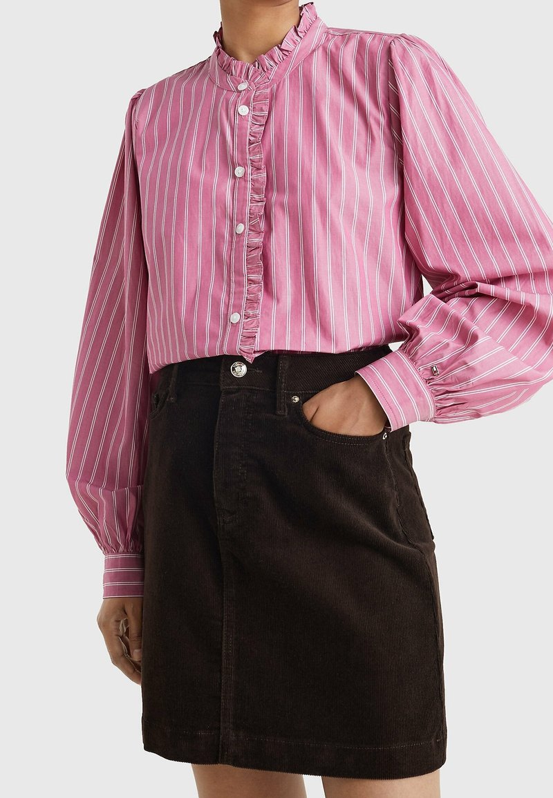 Femme portant un chemisier rayé rose avec des volants et des boutons, associé à une jupe en velours côtelé marron foncé taille haute avec des poches.