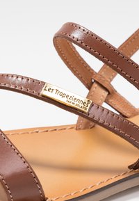 Sandalias de cuero marrón con correas delgadas que se cruzan. Una etiqueta de marca dorada dice "Les Tropéziennes par M. Belarbi." Plantilla de color beige claro con bordes cosidos.