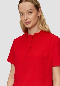 Rote Bluse mit kurzen Ärmeln, runder Ausschnitt und dezenten Falten. Glatte Stofftextur, taillierter Schnitt und neutraler Hintergrund.