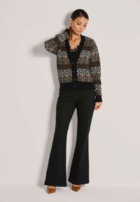 Frau in einem gemusterten Cardigan, schwarzem Spitzenoberteil, schwarzen Schlaghosen und braunen spitz zulaufenden Schuhen, die vor einer hellen Wand posiert.