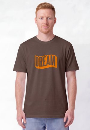 Jeune homme à la peau claire et aux cheveux roux portant un t-shirt marron avec un texte orange "DREAM", debout sur un fond blanc uni.