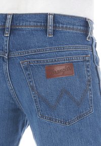 Jeans de mezclilla con una tela azul, corte recto, con una etiqueta de cuero de Wrangler y un distintivo bordado de los bolsillos traseros en un patrón de zigzag.