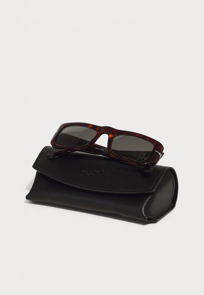 Gafas de sol de carey con lentes oscuros, descansando sobre un estuche de cuero negro. El estuche tiene una solapa curva y un detalle de logo en relieve.