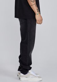 Jeans negros con corte recto y ligero desgaste, combinados con zapatillas blancas. El material parece suave con un diseño clásico de cinco bolsillos.