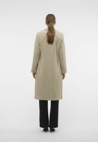 Beige lång kappa med en texturerad tyg och rak sk�ärning. Har en spetsig krage och långa ärmar, bärs över svarta byxor och skor.