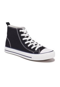 Zapatilla negra tipo botín de lona, con cordones blancos, puntera de goma y ribete blanco a lo largo de la suela.