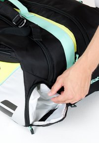 Hånd der åbner en sidelomme med lynlås på en sort og hvid sportsbag med lyseblå og gule detaljer på en hvid overflade.